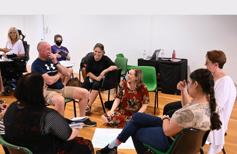 A group of men and women sitting on chairs in a circle, they are all talking and laughing. They are in a meeting room in what seems to be a workshop.