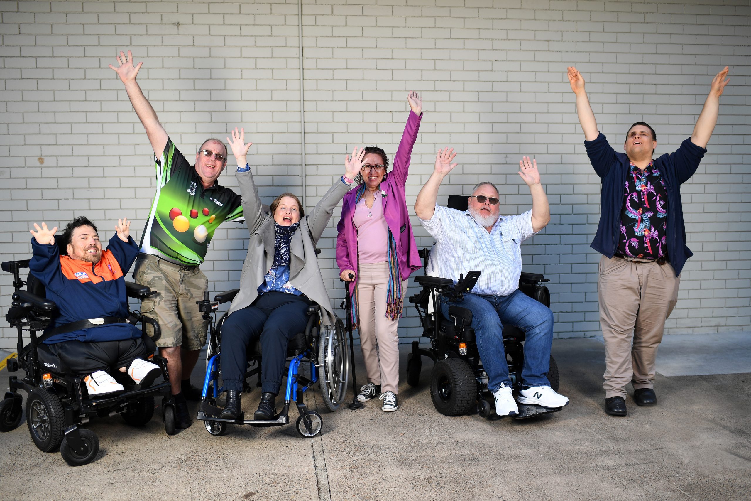 3 people are standing, 2 men and 1 woman, and 3 people in wheelchairs, 2 men and 1 woman, are all raising their arms in the air. They are standing in front of a white bricked wall.