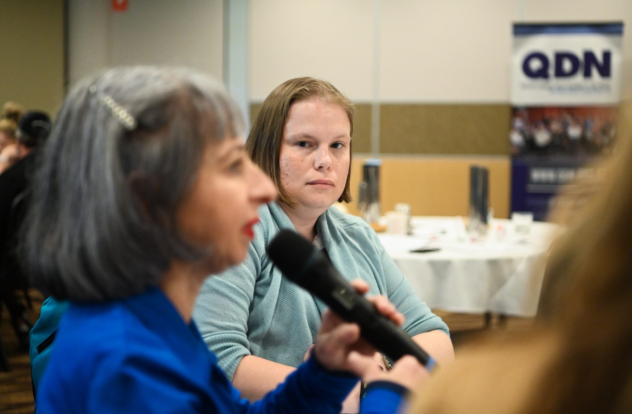 Two women, the one in the foreground is out of focus but she is holding a microphone. The woman in the background is in focus, she has short light brown hair and wearing a light blue long sleeved shirt.