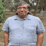 A man with glasses wearing a blue button up shirt smiling at the camera.