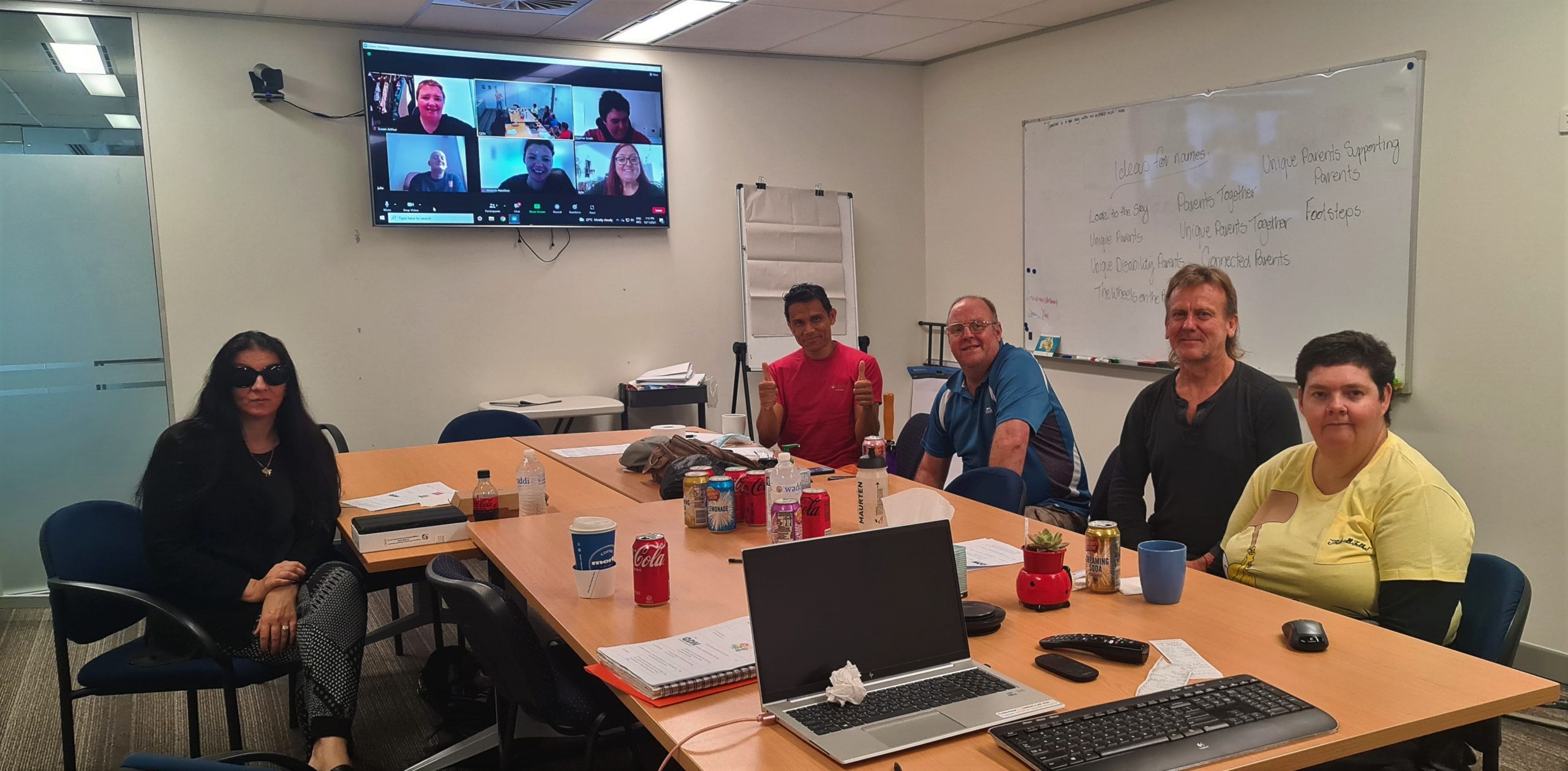 Five people sitting around a table in a board room, and a screen with 6 people on a zoom meeting on the back wall. They are all looking at the camera smiling.