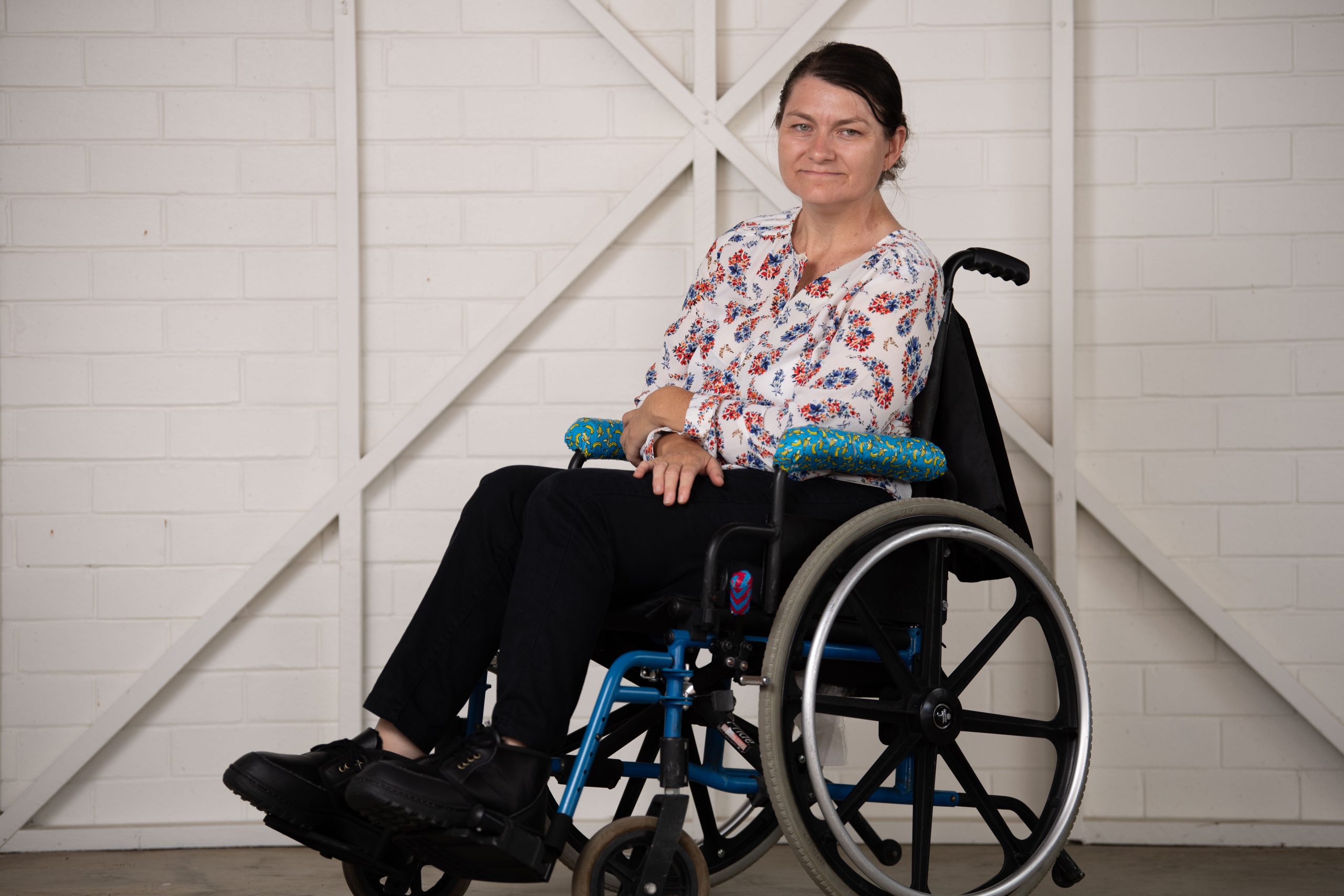 A woman with dark brown hair, wearing a white long sleeved patterned shirt with black pants and she is sitting in a wheelchair.