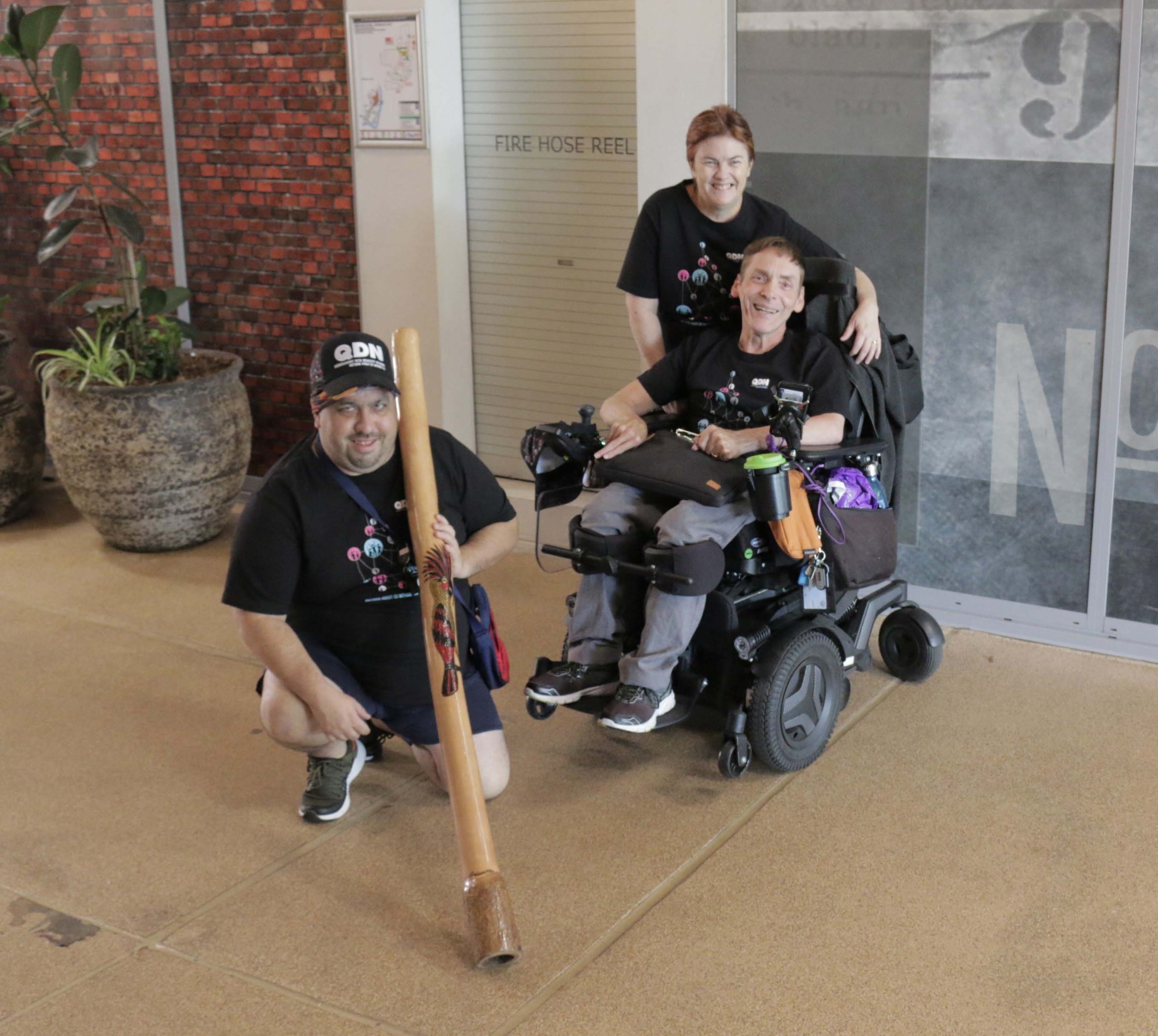 A man sitting in a wheelchair with a woman standing behind him and another man kneeling in front holding a didgeridoo. They are all wearing black QDN shirts.