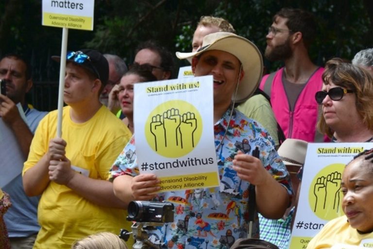A group of people protesting on the street, some people are standing, some are sitting, some are kneeling. Most people in the group are wearing yellow t-shirts. Multiple people in the group are holding posters. The posters each have an illustration of raised fists within a yellow circle and text that reads ‘Stand with us‘, #StandWithus, Queensland Disability Advocacy Alliance”. Brendon Donohue is standing in the crowd, smiling, and facing the camera, he is wearing a blue patterned shirt and is holding one of the posters described above.