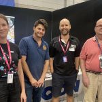 A photo of four people standing looking at the camera smiling. Two of the people are QDN staff members wearing QDN t shirts, the other two people are QDN members. They are standing in front of the QDN stall at the Brisbane Disability Connection expo.