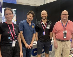 A photo of four people standing looking at the camera smiling. Two of the people are QDN staff members wearing QDN t shirts, the other two people are QDN members. They are standing in front of the QDN stall at the Brisbane Disability Connection expo.