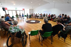 A circle of people with disabilities all facing each other in a room, listening to one person speak.