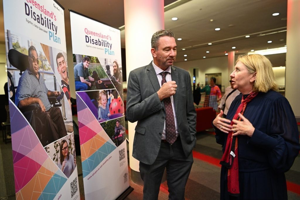 A man and a woman who are standing in a large grand looking room. They are standing next to some marketing pull up banners promoting Australia's Disability Plan. They appear to be mid conversation. The women is wearing a blue dress, red patterned scarf and has short blond hair that sits above her shoulders. The man is wearing grey suit, white shirt, and dark red tire, with dark short cropped hair.