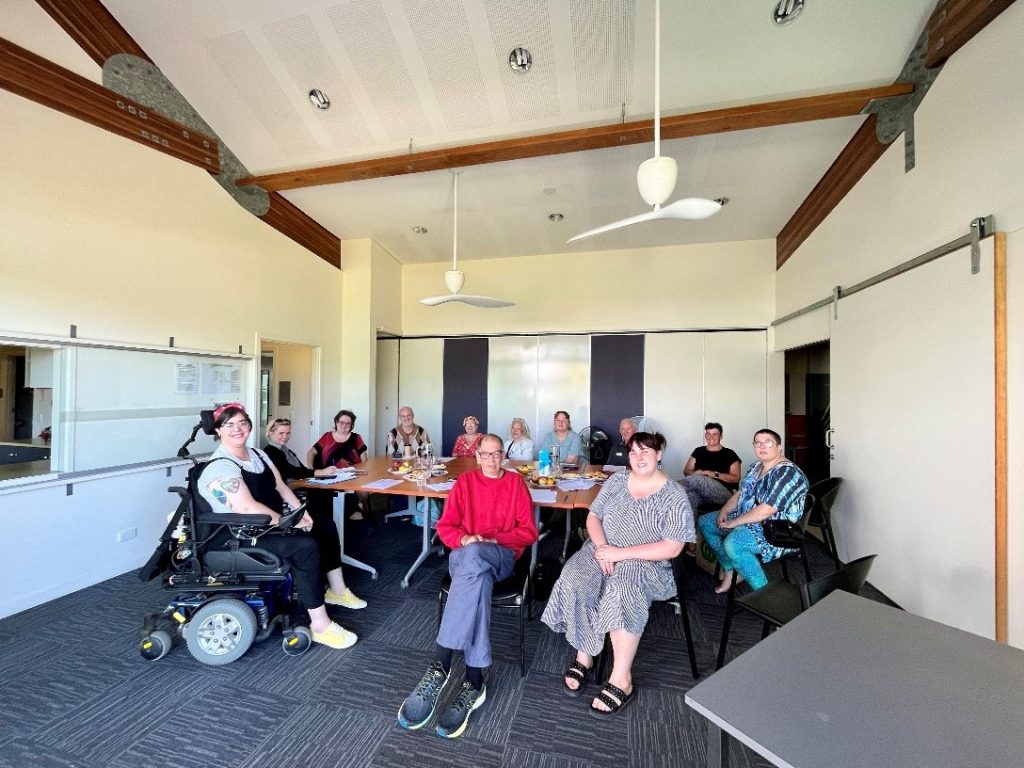 A group of 12 people in a large, bright room with tall ceilings.. They are all sitting around a large table with paper and glasses of water. One person is using a power wheelchair. They are all looking towards the camera smiling.