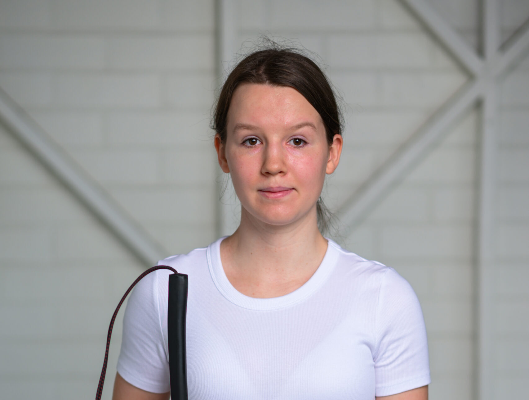 A woman with dark brown hair, holding a white cane, is wearing a white T-shirt and smiling in front of a plain white background.