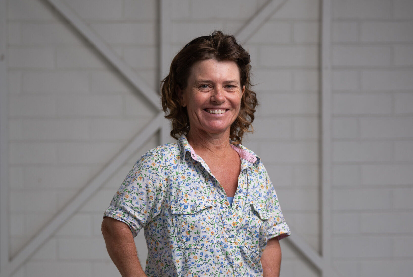 A woman with shoulder-length brown hair, wearing a white floral T-shirt, is smiling in front of a plain light-colored background.