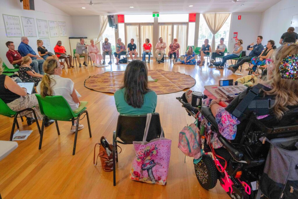 A circle of people with disabilities all facing each other in a room, listening to one person speak.