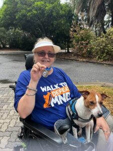 Image: Carmen Jen, a QDN member and 2024 Emerging Leader, sits in her wheelchair holding her MND Cornflower Spirit Award. A dog sits beside her.