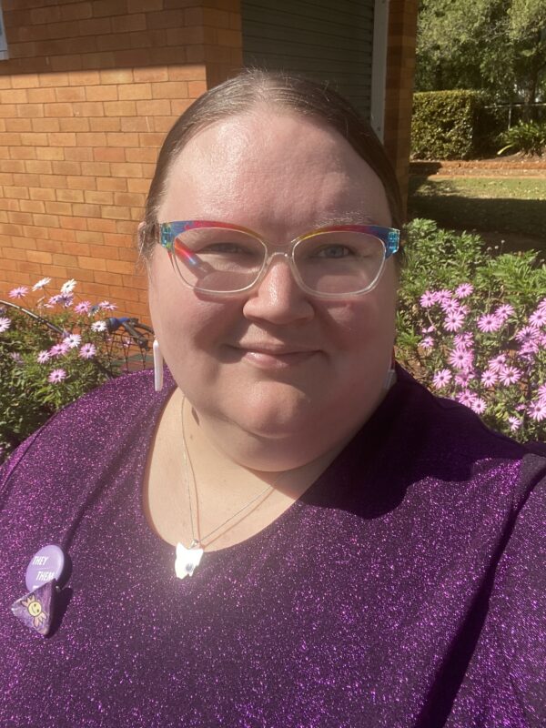 A woman wearing a sparkly purple top and rainbow-framed glasses, standing in front of a garden with pink flowers. They have a 'they/them' pronoun badge and other pins on their top. A brick wall and greenery are visible in the background.