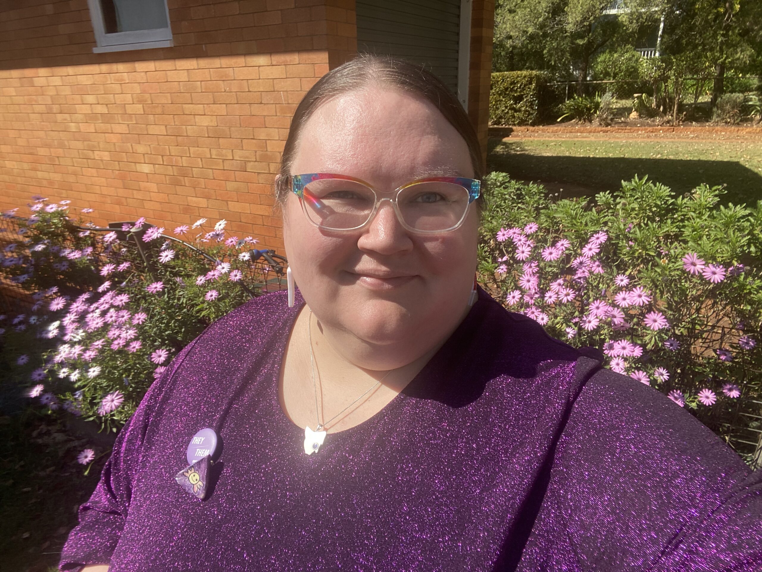 A person wearing a sparkly purple top and rainbow-framed glasses, standing in front of a garden with pink flowers. They have a 'they/them' pronoun badge and other pins on their top. A brick wall and greenery are visible in the background.