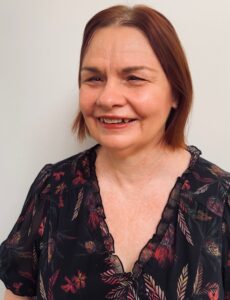 A person with shoulder-length brown hair, wearing a dark floral blouse, smiling in front of a plain light-colored background.