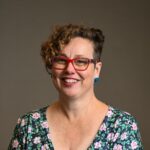 Woman with short brown curly hair wearing a floral dress and red glasses