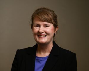 Woman with short dark hair wearing a purple shirt and a black blazer.
