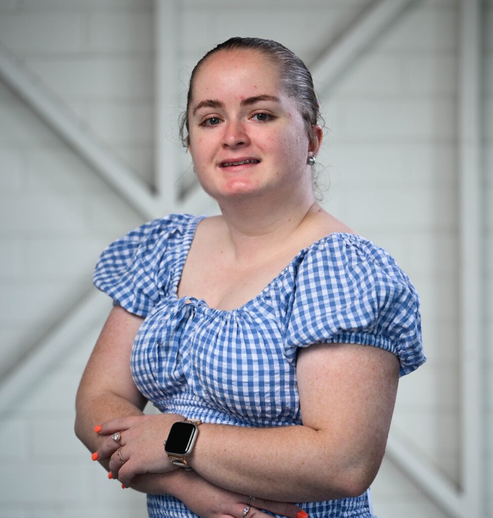 A woman standing and smiling at the camera. She has light brown hair tied back into a ponytail. She is wearing a light blue and white gingham patterned dress with capped sleeves.
