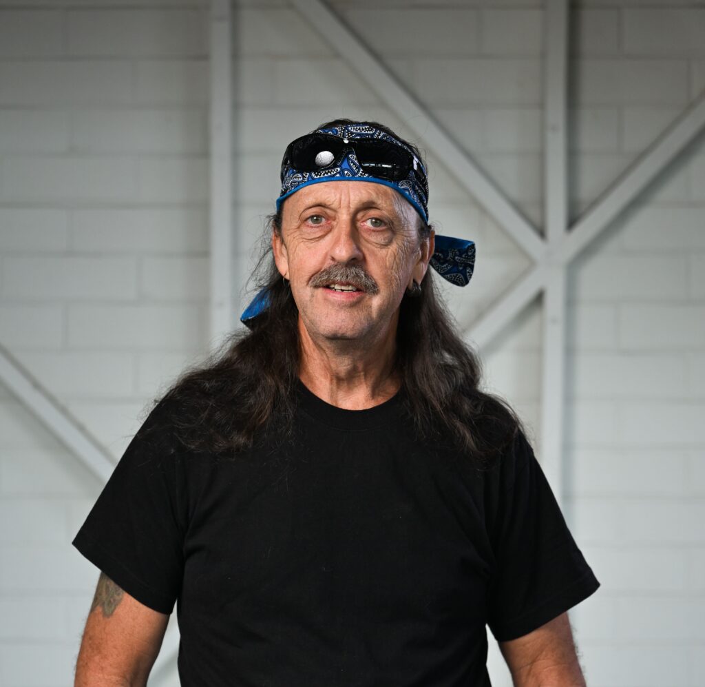 A man standing, looking at the camera with a big smile on his face. He has his thumbs hooked into his front pockets. He has long dark hair and a mustache. He is wearing a blue paisley bandana and sunglasses on his head, a black t-shirt and blue denim shorts with a black belt. He has two tattoos on his right forearm, and a tattoo on his wrist and a tattoo on his left forearm. He is wearing a prosthetic left leg just above his knee.