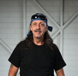 A man standing, looking at the camera with a big smile on his face. He has his thumbs hooked into his front pockets. He has long dark hair and a mustache. He is wearing a blue paisley bandana and sunglasses on his head, a black t-shirt and blue denim shorts with a black belt. He has two tattoos on his right forearm, and a tattoo on his wrist and a tattoo on his left forearm. He is wearing a prosthetic left leg just above his knee. 