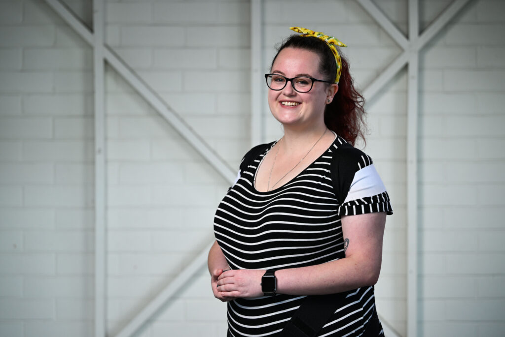 A woman with glasses standing in front of the camera smiling. She has long, dark wavy hair tied back into a ponytail with a yellow patterned head scarf. She is wearing a black and white striped t-shirt and a black harness.