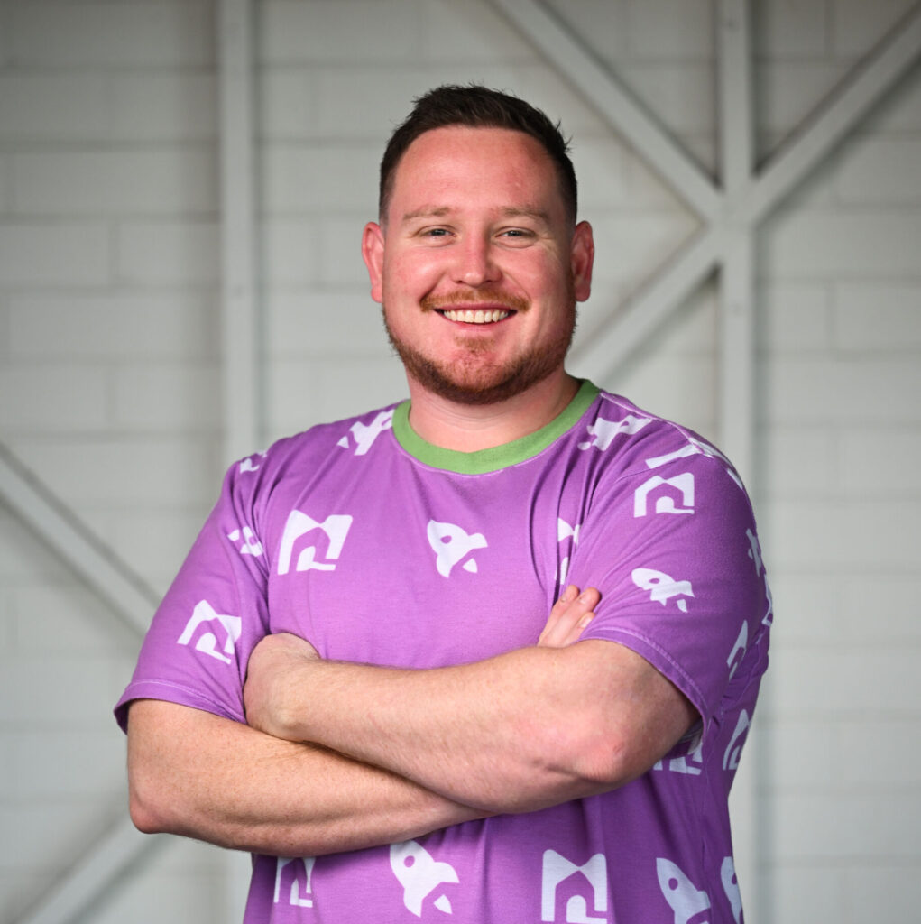 A man smiling, looking at the camera with his arms crossed. He has short dark red hair and a short beard. He is wearing a purple top with pictures of small rockets and a green collar
