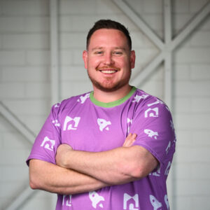 A man smiling, looking at the camera with his arms crossed. He has short dark red hair and a short beard. He is wearing a purple top with pictures of small rockets and a green collar