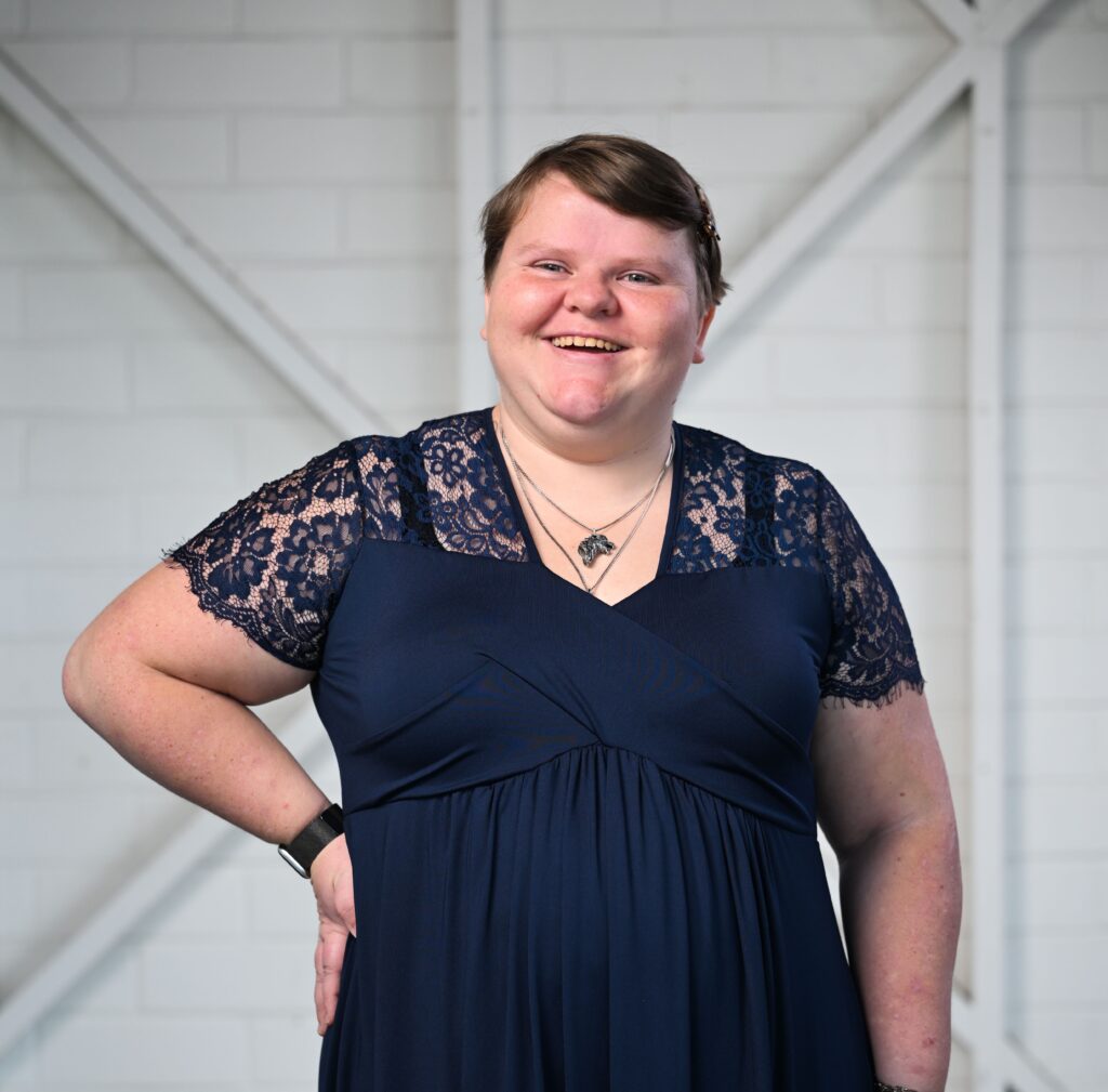 A woman standing smiling and looking at the camera. She has short, brown hair and is wearing a dark blue dress with short, lace sleeves.