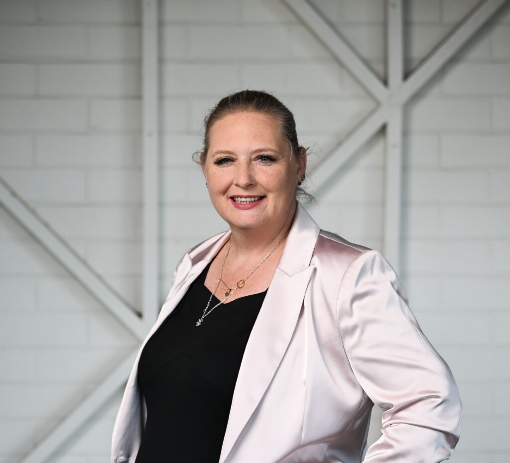 Woman facing side on and smiling at the camera. She is wearing a beige satin blazer and a black shirt.