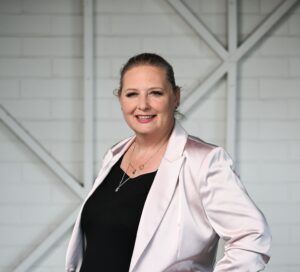 A woman standing with her arms crossed, smiling at the camera. She has blonde hair tied back into a ponytail. She is wearing a dark top with a silky pale pink blazer.