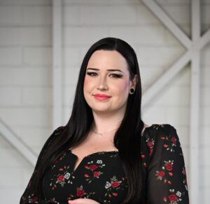 A woman standing and smiling at the camera. She has long, black hair and is wearing a black long-sleeved dress with a red floral pattern.