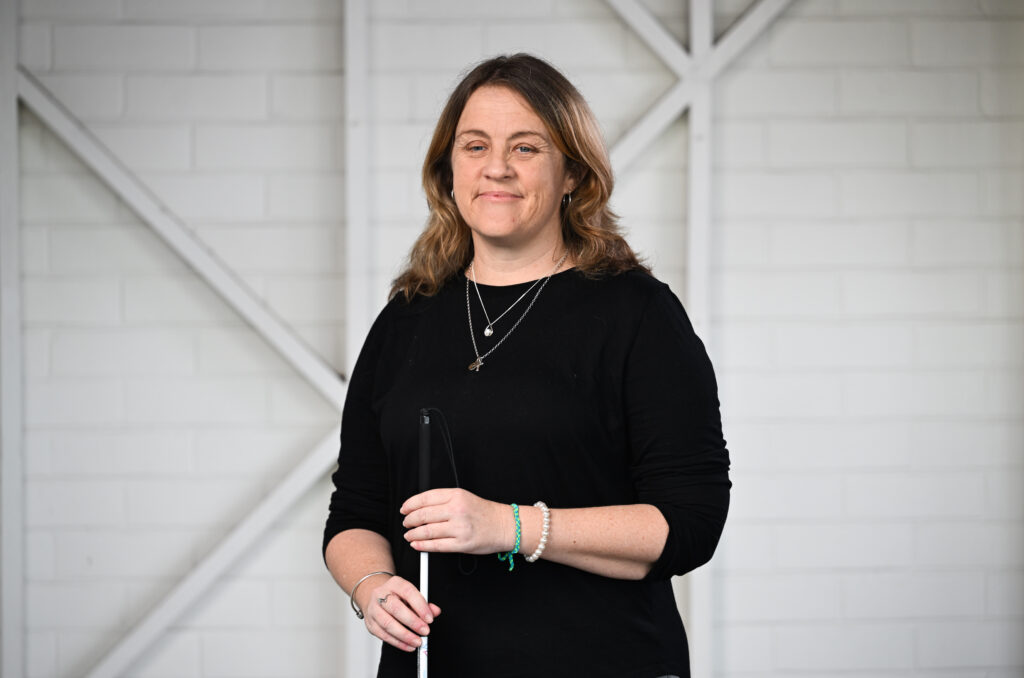 A woman smiling and looking at the camera. She has shoulder length brown hair. She is wearing a black top, grey pants and is holding a silver cane.