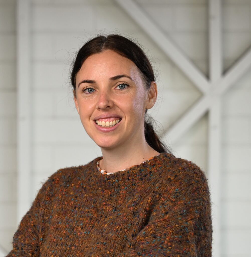 A woman standing in front of the camera with her hands in her front pockets, smiling at the camera. She has dark hair tied back into a low ponytail. She is wearing a brown short-sleeved jumper with light coloured pants.
