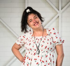 A woman looking at the camera with her head tilted to the left and a big smile. She has long dark hair tied into a high ponytail. She is wearing a white dress with a pattern of red cherries with green stems throughout.