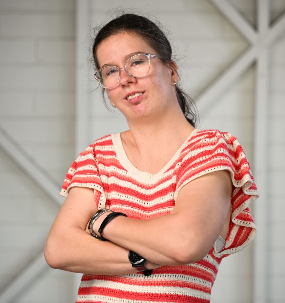 A woman with glasses standing in front of the camera, smiling. She has her arms crossed and her head tilted to the left. She is wearing a terracotta and white stripped dress with short flowy sleeves.