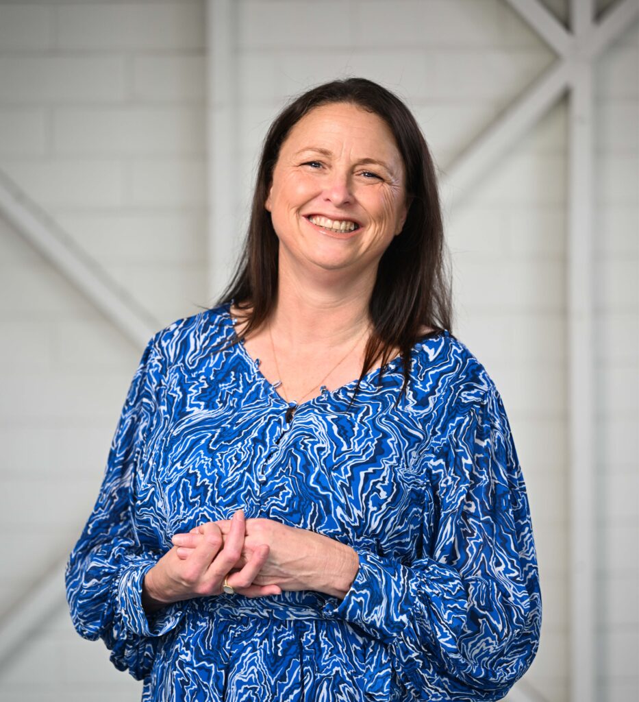A woman standing and smiling at the camera. She has dark, shoulder length hair and is wearing a blue swirly-patterned, long-sleeve dress.