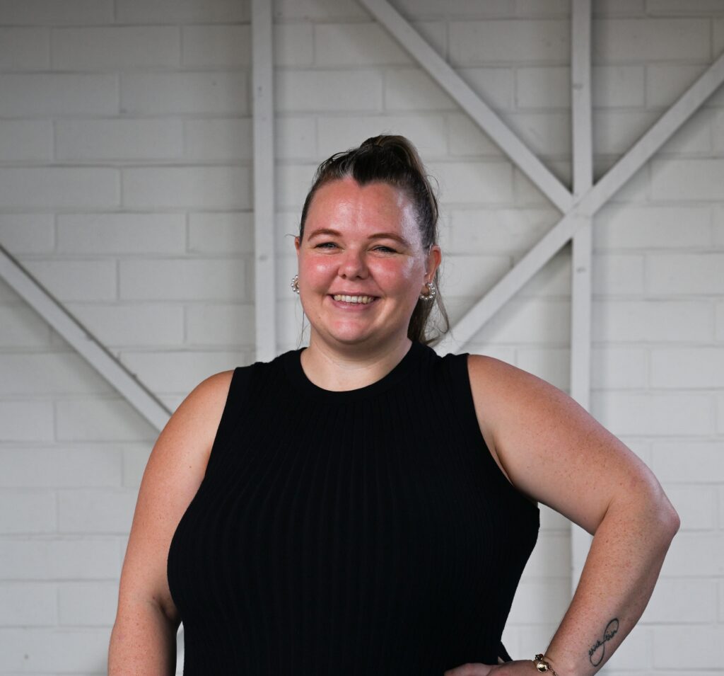 A woman standing and smiling at the camera, with her hand on her left hip. She has dark hair pulled back into a high ponytail. She is wearing a black sleeveless top with black pants.