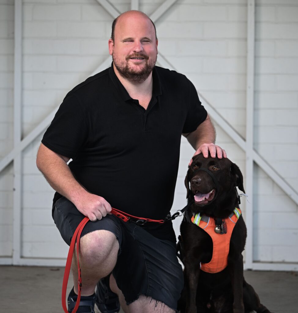 A man crouching and smiling at the camera. He is wearing a black polo shirt, dark denim shorts and black trainers. He is holding the lead to an assistance dog who has dark fur and is wearing an orange harness.