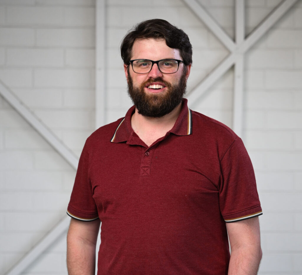 A man standing with his hands in his pockets, smiling at the camera. He has short dark hair, medium-length dark beard and is wearing glasses. He is wearing a maroon coloured polo shirt with a thin white strip on the sleeve cuffs and collar and brown pants.
