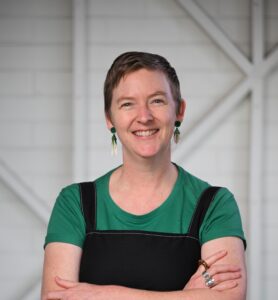 A woman standing with her arms crossed and smiling at the camera. She has short, light brown hair. She is wearing a black, thin strapped dress over an emerald, green t-shirt and green earrings. 