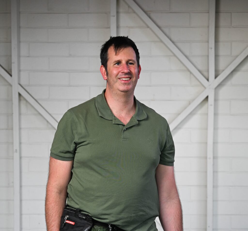 A man standing and smiling at the camera. He has short, dark hair and is wearing a green polo shirt, black jeans and blue and orange trainers. He is holding the lead to a black assistance dog who is wearing a blue harness. He is wearing a black belt bag around his waist.