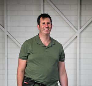 A man standing and smiling at the camera. He has short, dark hair and is wearing a green polo shirt, black jeans and blue and orange trainers. He is holding the lead to a black assistance dog who is wearing a blue harness. He is wearing a black belt bag around his waist.