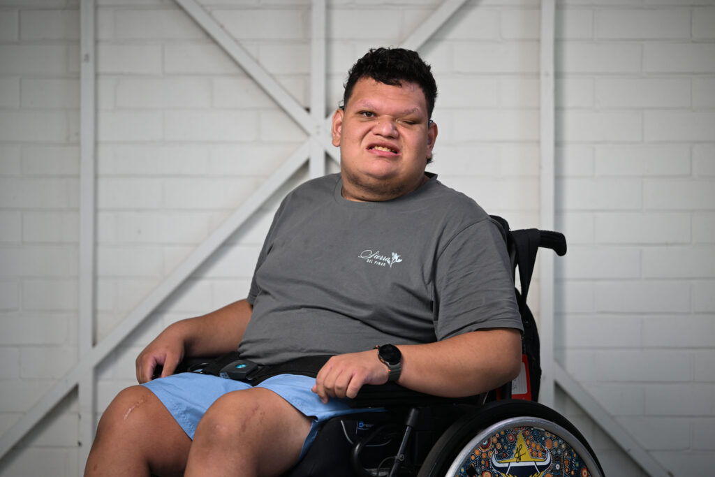 A man in a wheelchair facing and smiling at the camera. He has short dark hair and is wearing a grey t-shirt, blue shorts and black sandals. His back left wheelchair wheel is decorated with North Queensland Cowboys logos.