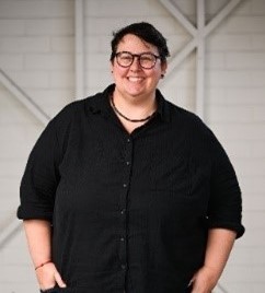 A woman standing in front of the camera with her hands in her front pockets, smiling. She has short dark hair and is wearing glasses. She is wearing a black button up shirt with dark blue jeans.