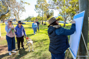 In a park, a woman in a puffer vest and Akubra hat is writing on a whiteboard. Two women are standing close by, one with a guide dog, and one with a cane.