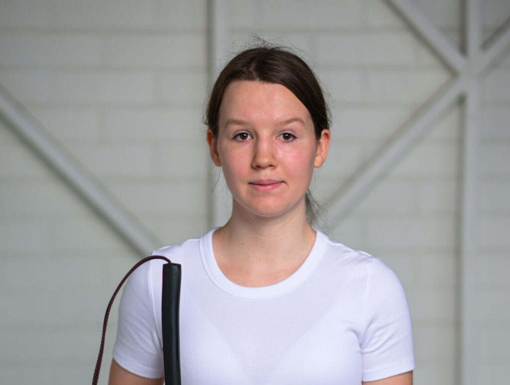A woman in a white shirt facing the camera, she is holding a cane. 