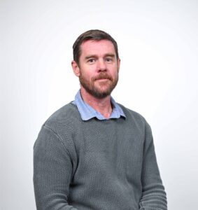 man with a beard looking at the camera, wearing a blue shirt and grey jumper.