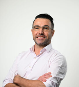 Man wearing a pink shirt looking at the camera, he also is wearing glasses.