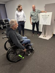 A man in a wheelchair wearing blue jeans and a grey jumper looking at an eye chart with 2 other men observing